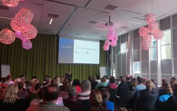 Foto van een ruimte met een beamerscherm, presentator en zittend publiek