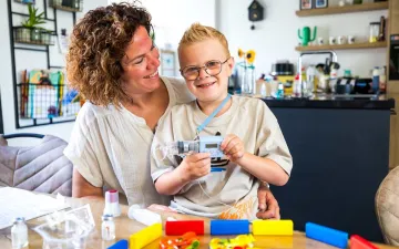 Moeder en zoon zitten aan de keukentafel. De jongen houdt het infuus met plasmamedicijnen vast en lacht. 