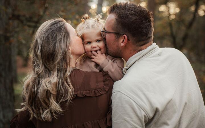 Foto van meisje in de armen van haar moeder die tussen haar ouders doorkijkt de camera in. 
