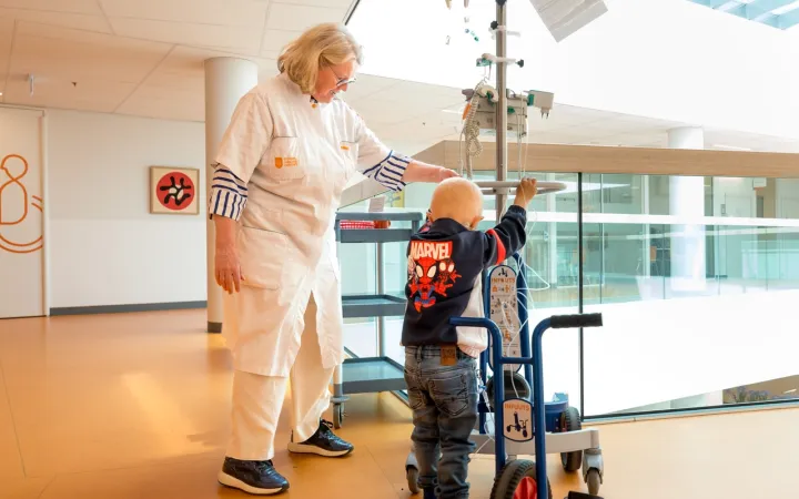 Foto van kinderoncoloog en jonge patient in de ziekenhuisgang. Samen duwen ze een fuuts aan, een fiets met infuus. 