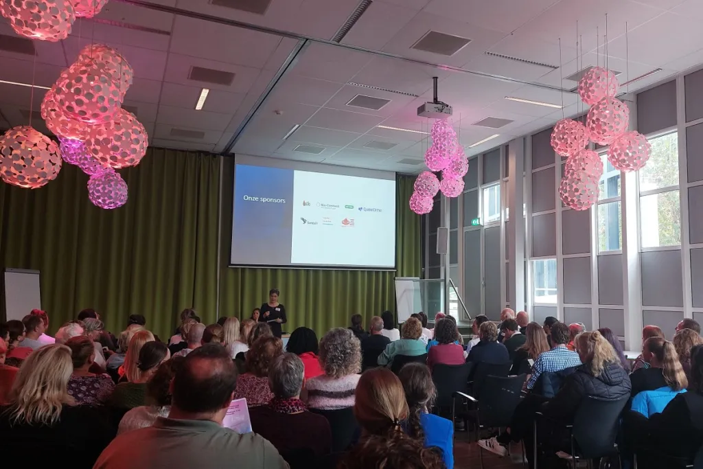 Foto van een ruimte met een beamerscherm, presentator en zittend publiek