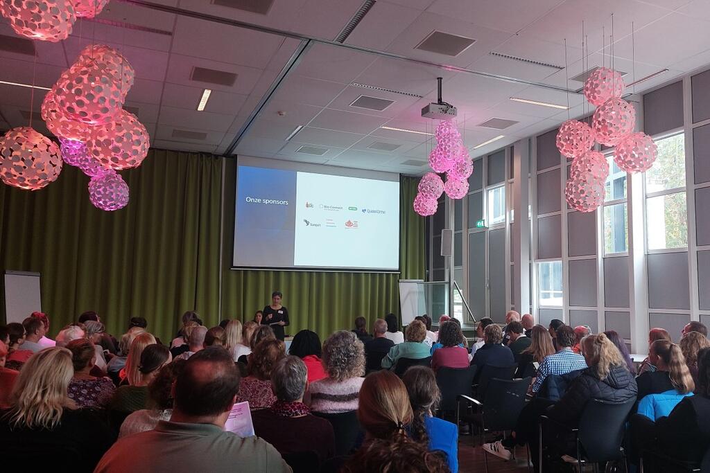 Foto van een ruimte met een beamerscherm, presentator en zittend publiek