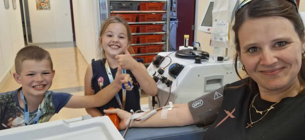 Foto van Marielle tijdens het doneren van plasma. De kinderen staan lachend naast de stoel. 