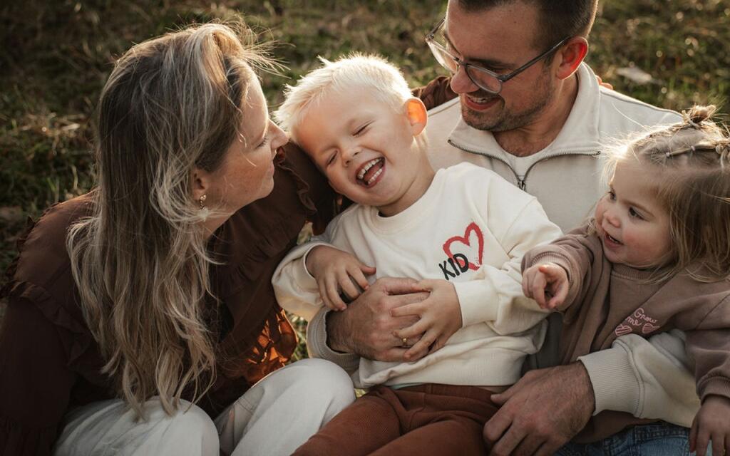 Foto van moeder, vader en twee kinderen zittend in het gras. Het jongetje lacht en het meisje wijst naar de camera.