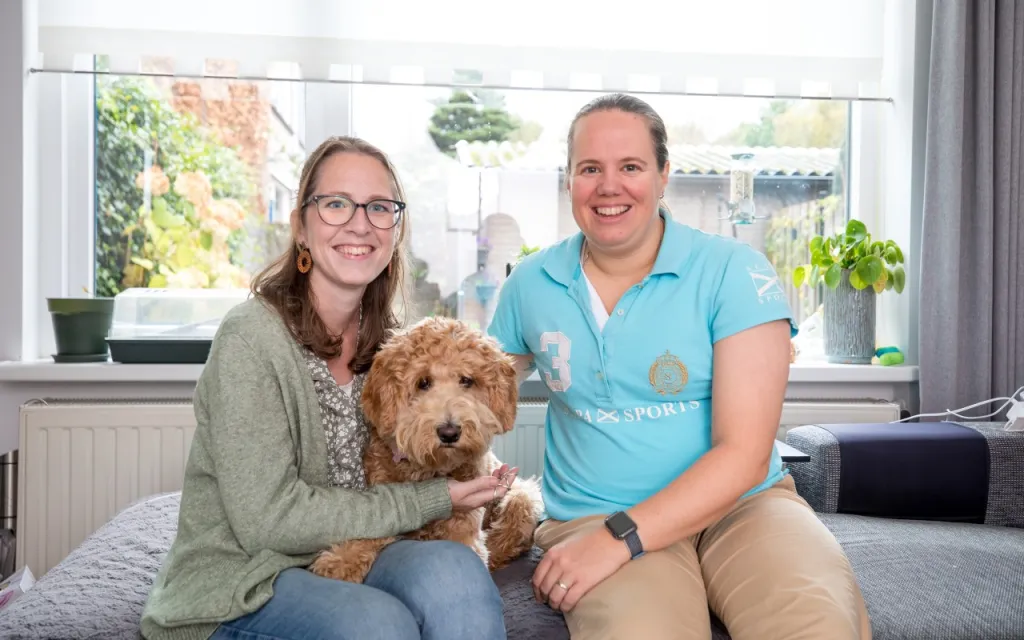 foto van Nicky (links) en Irene (rechts) op een bank in een woonkamer. Ertussenin zit een hond