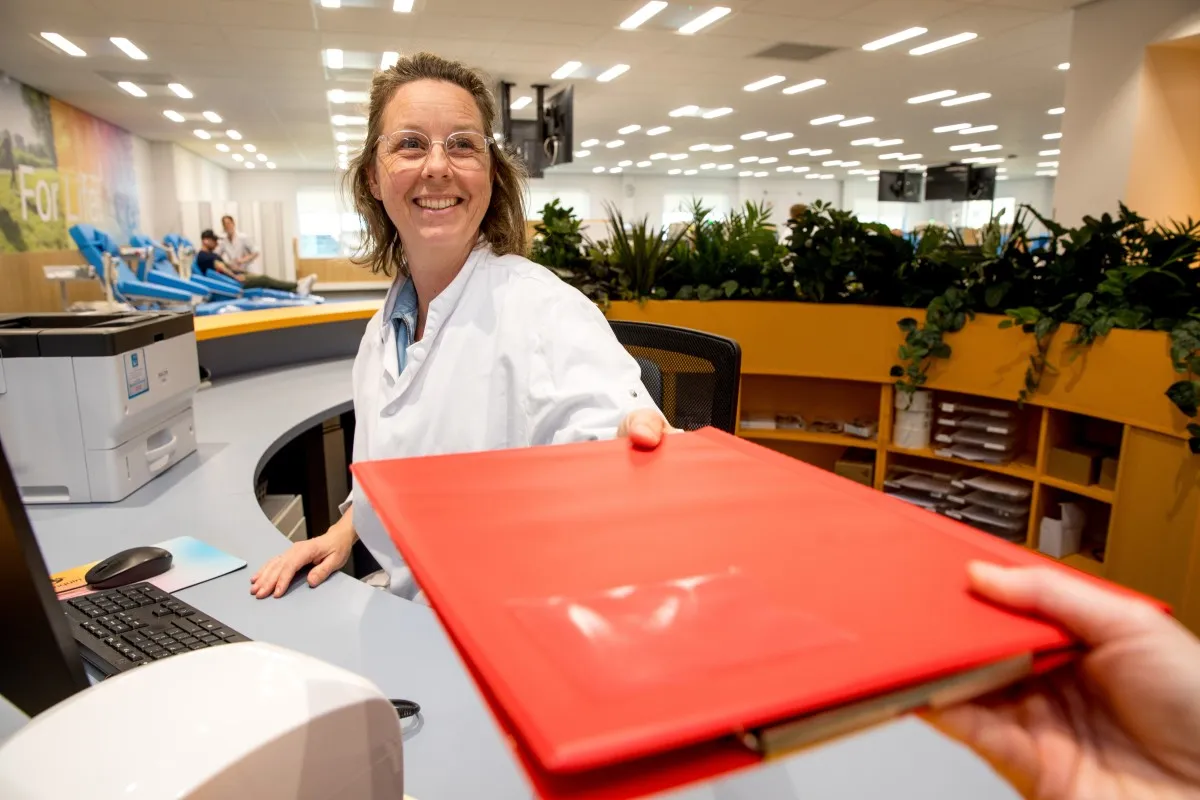 Medewerker aan de balie overhandigd lachend een rode map met de medische vragenlijst